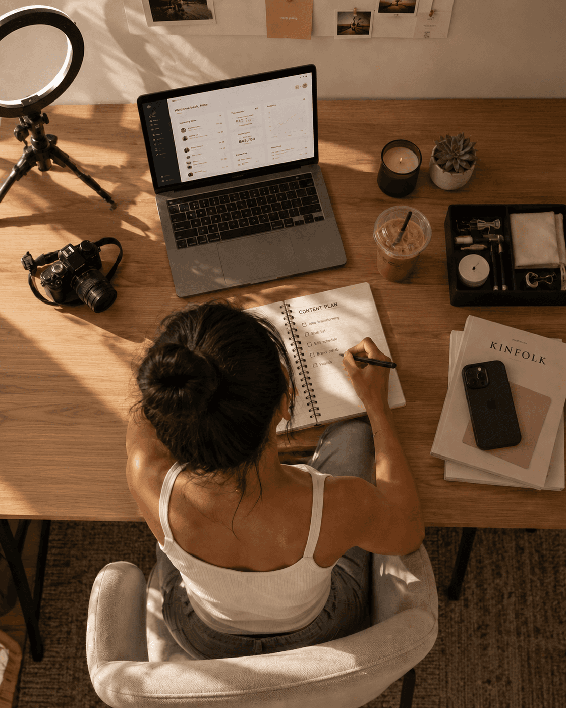 A creator at her desk seen from above, writing a content plan in a notebook beside a laptop, a camera and a ring light.